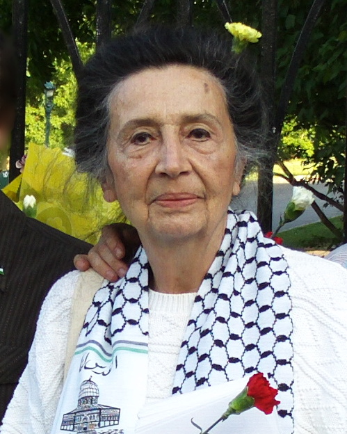 Marie-Claude regarde sereinement et directement la caméra, devant de la verdure près des Plaines d'Abraham à Québec. Elle porte un foulard blanc avec un motif noir, symbol des Palestinien-nes et tient aussi une rose dans ses mains.
