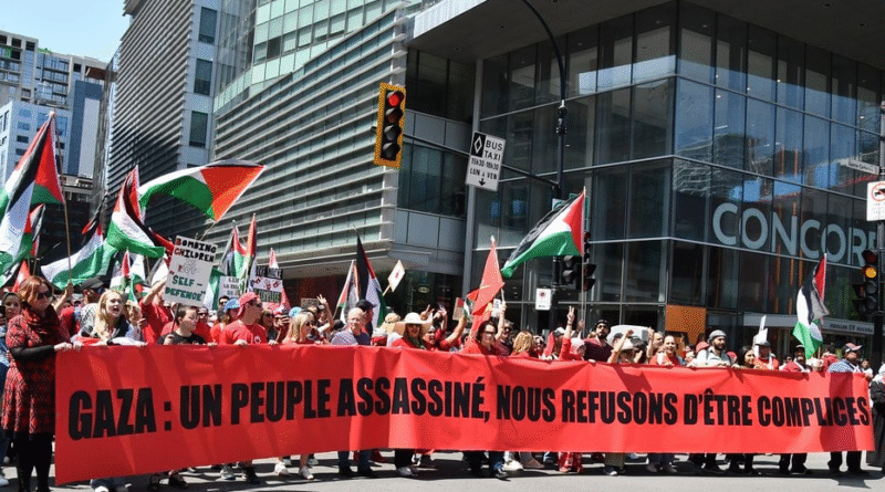 Photo du devant d'une manif prenant une large voie et la bannière de front est très longue et se lit: Gaza: un peuple assassiné, nous refusons d'être complices. Plusieurs drapeaux palestiniens. Source: urgencepalestine.quebec